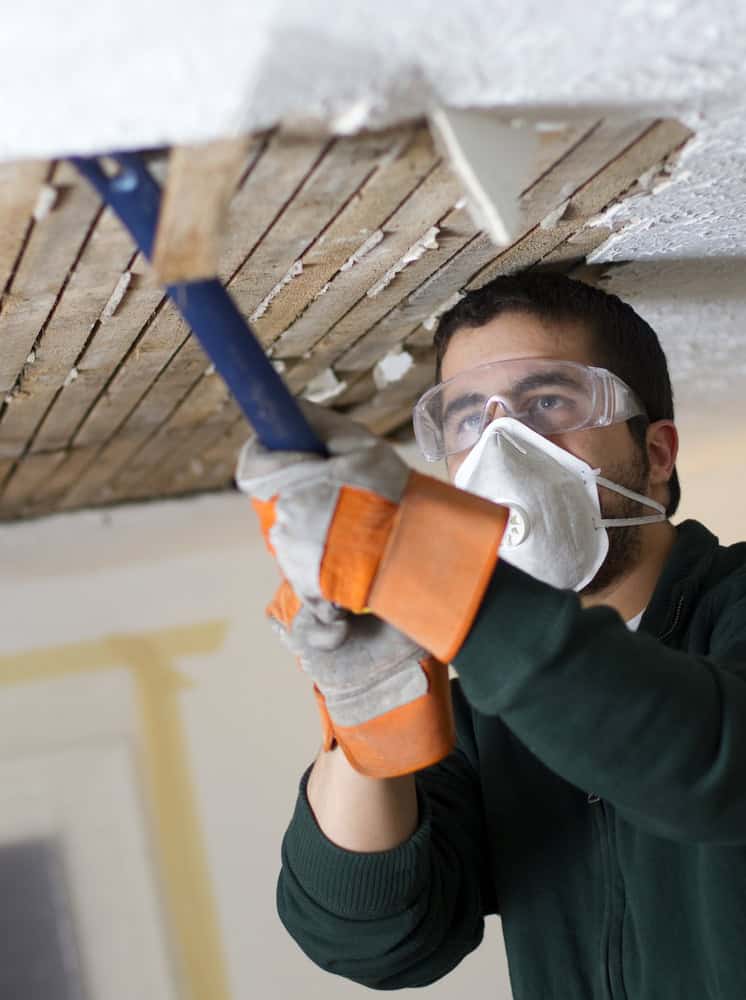 A person wearing protective goggles, a mask, and gloves uses a crowbar to remove plaster from a ceiling, revealing wooden lath underneath. Focused on water damage restoration in Palm Beach County, the individual ensures safety precautions are observed during the intricate task.
