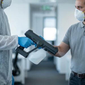 Two people wearing masks and gloves, one in protective coveralls, are exchanging a disinfectant spray device in an office setting.