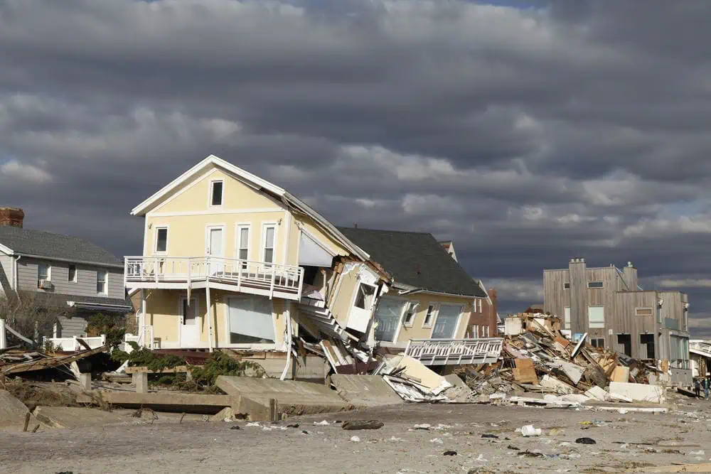 A yellow house is severely damaged and leaning, debris scattered around—a testament to the storm's aftermath. Nearby houses in Palm Beach County stand under a cloudy sky, highlighting the urgent need for storm damage restoration and water mitigation services.