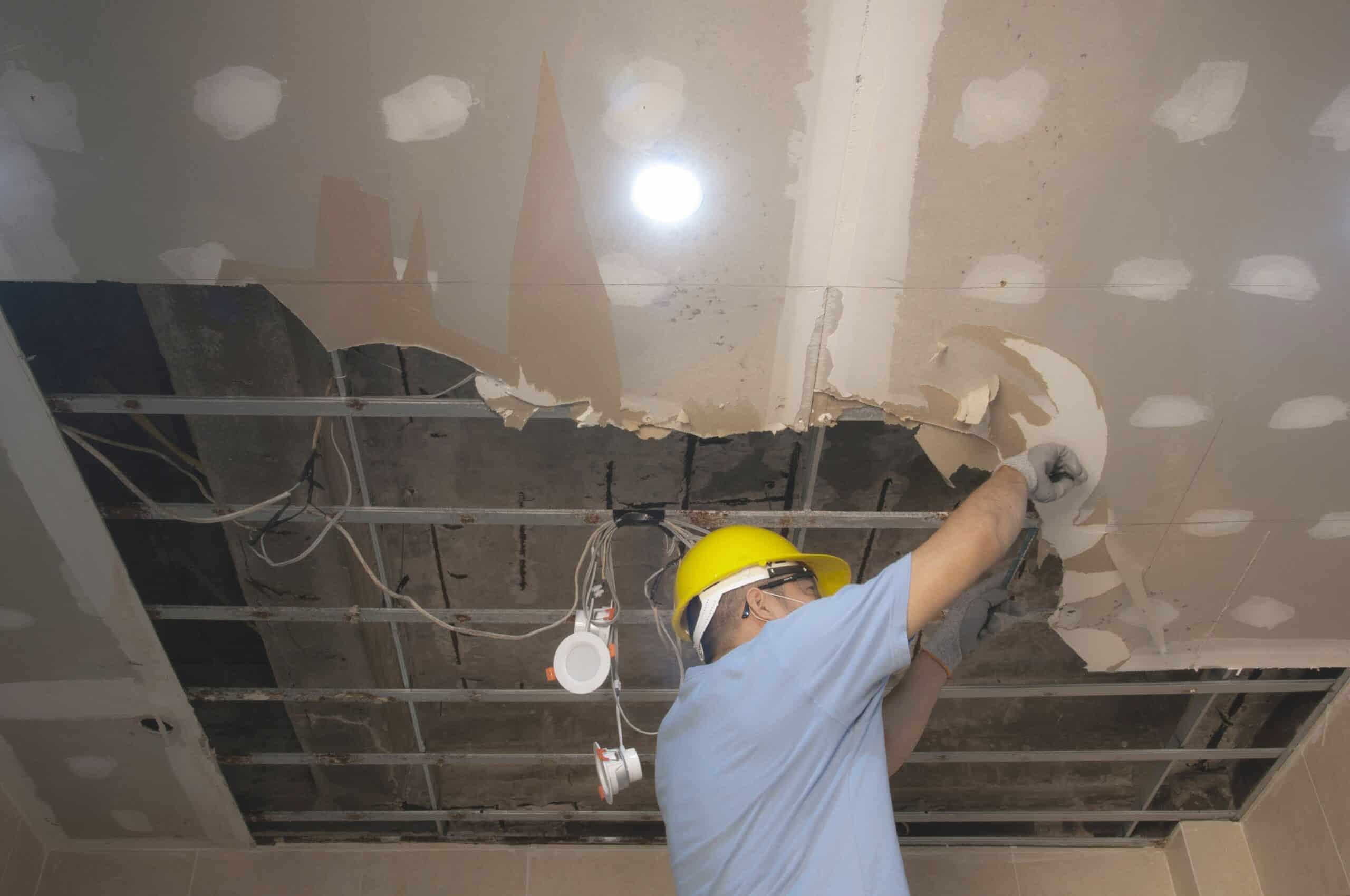 A worker in a blue shirt and yellow hard hat is repairing a damaged ceiling, removing drywall caused by storm damage. The ceiling has a large hole exposing wiring and beams, highlighting the need for storm damage restoration services in Palm Beach County. The room is lit with bright ceiling lights.
