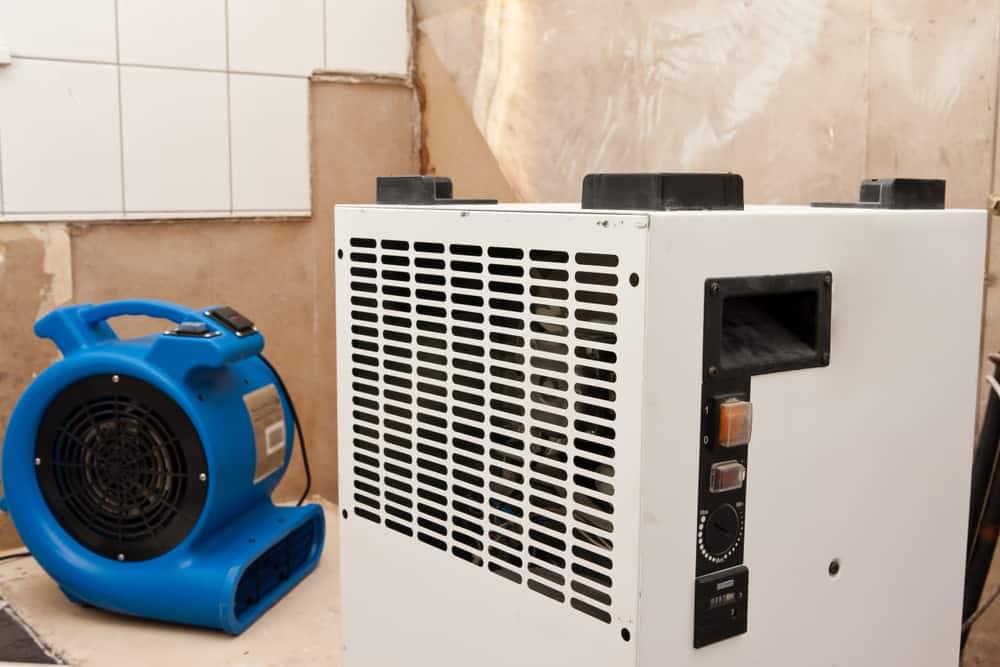 A white dehumidifier and a blue air mover are strategically positioned in a room featuring exposed drywall and tiled walls, essential for drying and ventilation. This setup indicates ongoing water mitigation services in Palm Beach County, crucial for effective storm damage restoration.