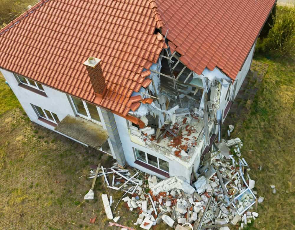 Aerial view of a house with significant structural damage. The roof and part of the wall have collapsed, exposing the interior. Debris, including bricks and wooden beams, are scattered around the foundation. The house has a red tiled roof.