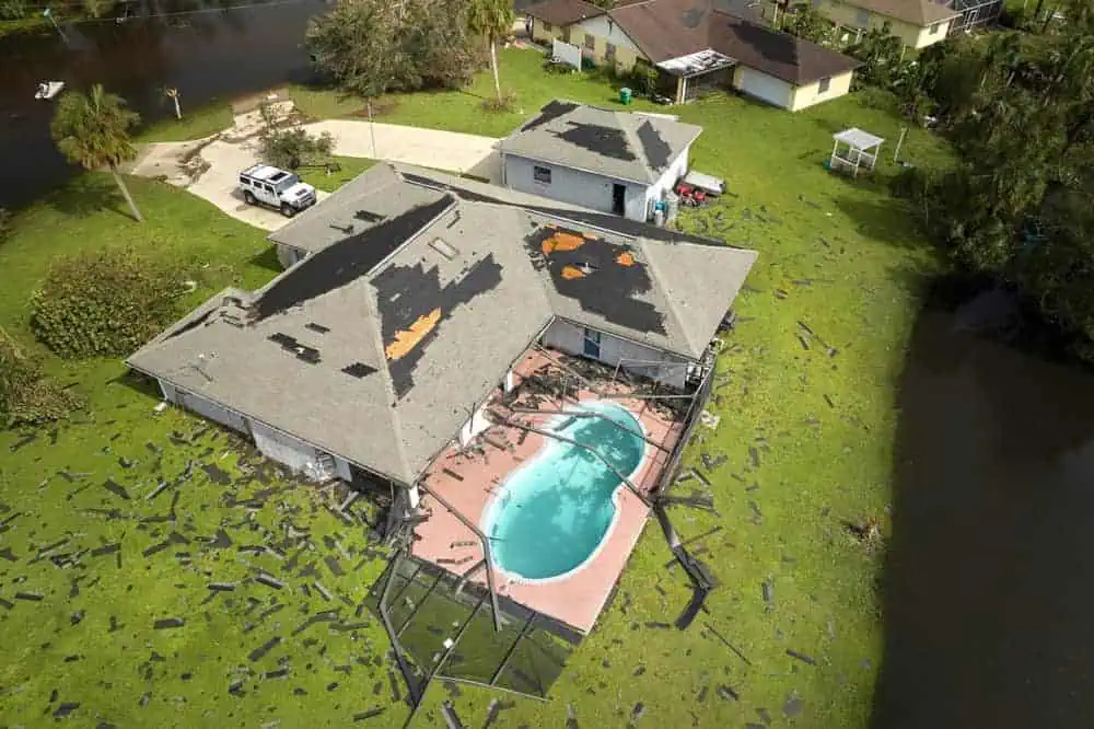 Aerial view of a house with a damaged roof, missing shingles scattered on the ground. A small pool is in the backyard amid signs of storm damage. Water surrounds the area, hinting at recent flooding, making storm damage restoration in Palm Beach County essential. Nearby trees and homes are visible.