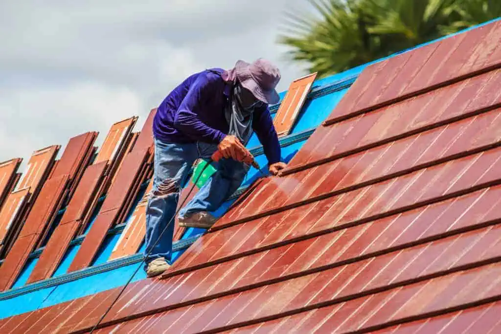 A worker in a purple shirt and hat carefully installs reddish-brown tiles on a rooftop, with partly cloudy skies in the background. Palm tree fronds are visible, suggesting a warm climate.