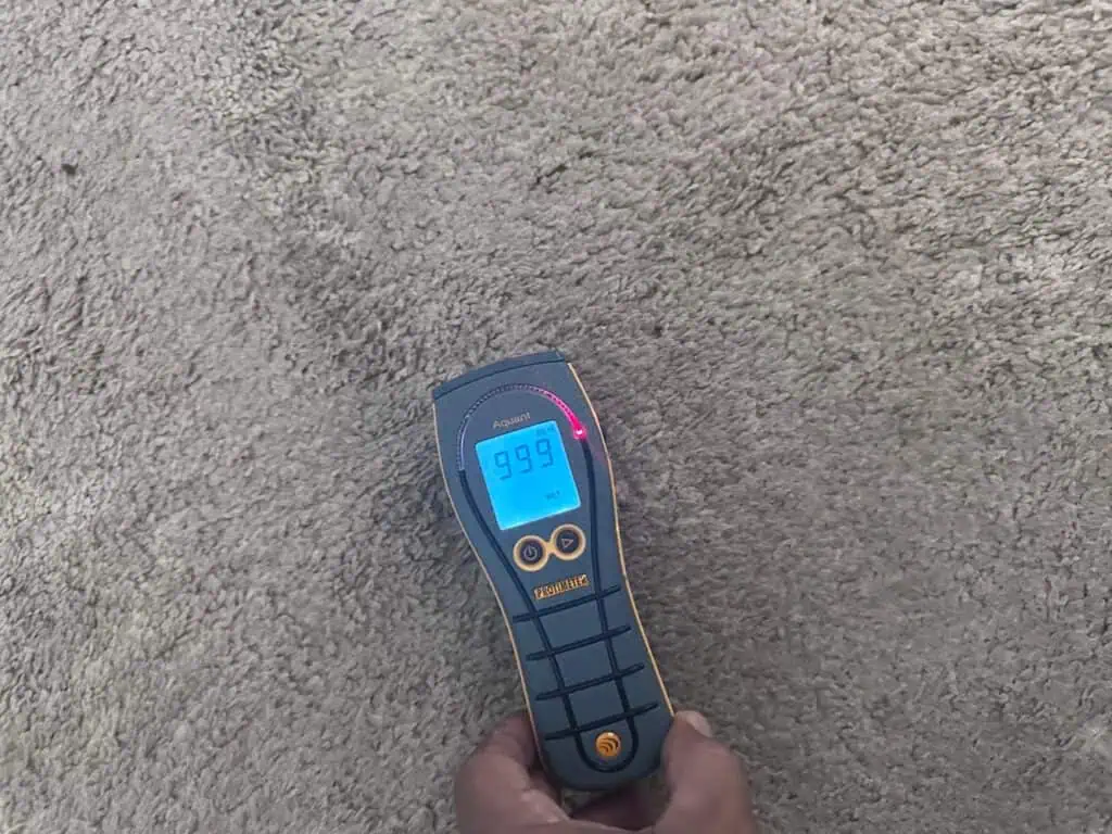 A handheld moisture meter displaying a reading of 9.9% on a digital screen is placed on a beige carpet. A red indicator light is visible above the screen, and a hand is holding the device.